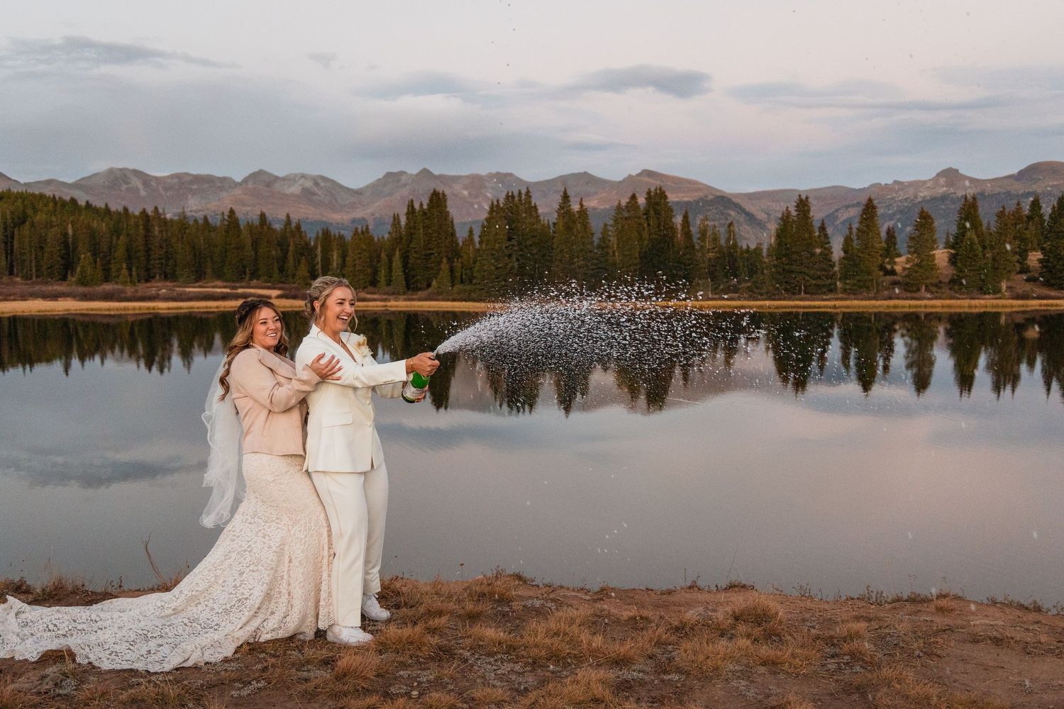 A couple in wedding attire stands by a serene mountain lake at sunset with reflective waters and pine trees in the background.