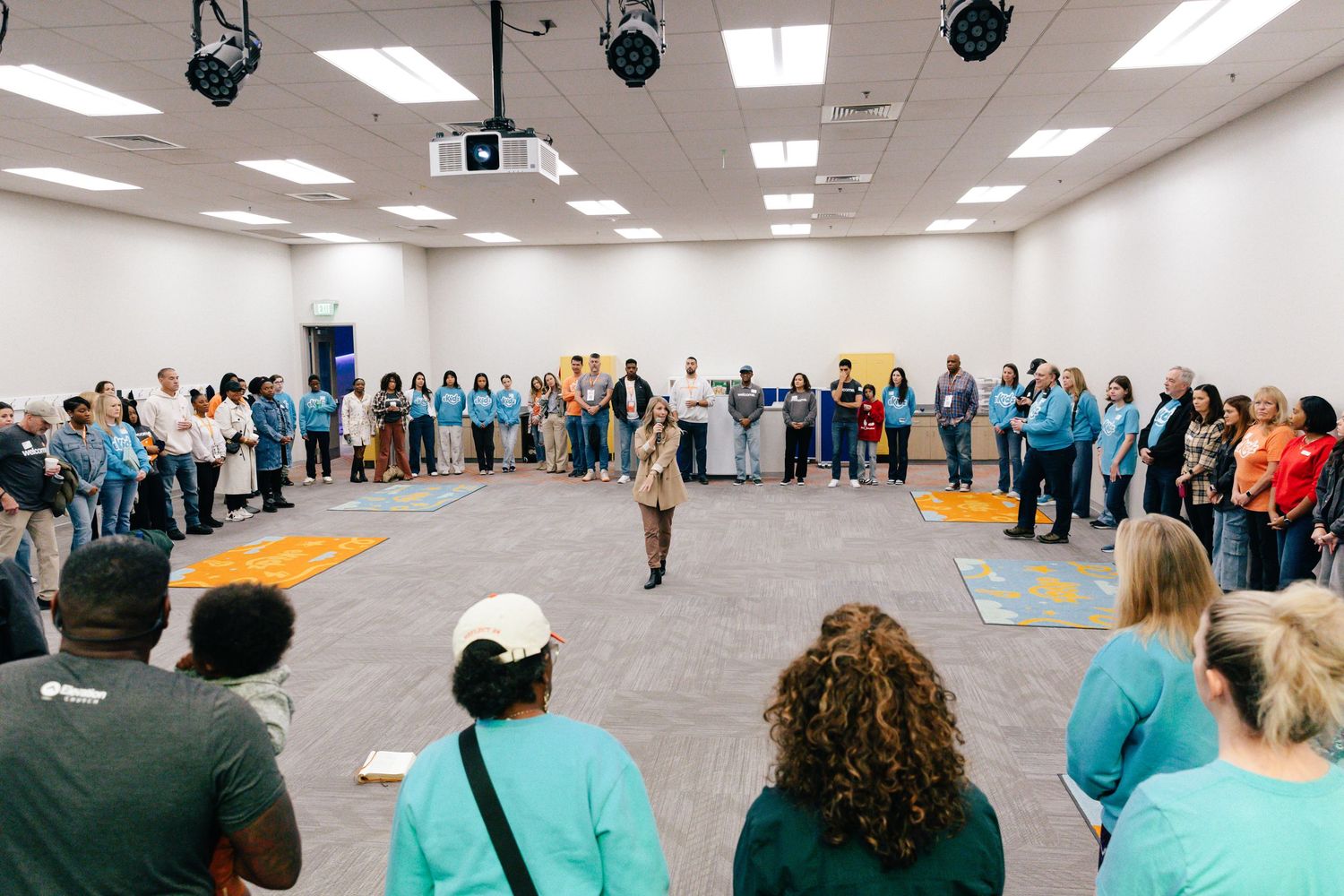 A large group of people stand in a circle during an indoor community gathering event in a bright room.