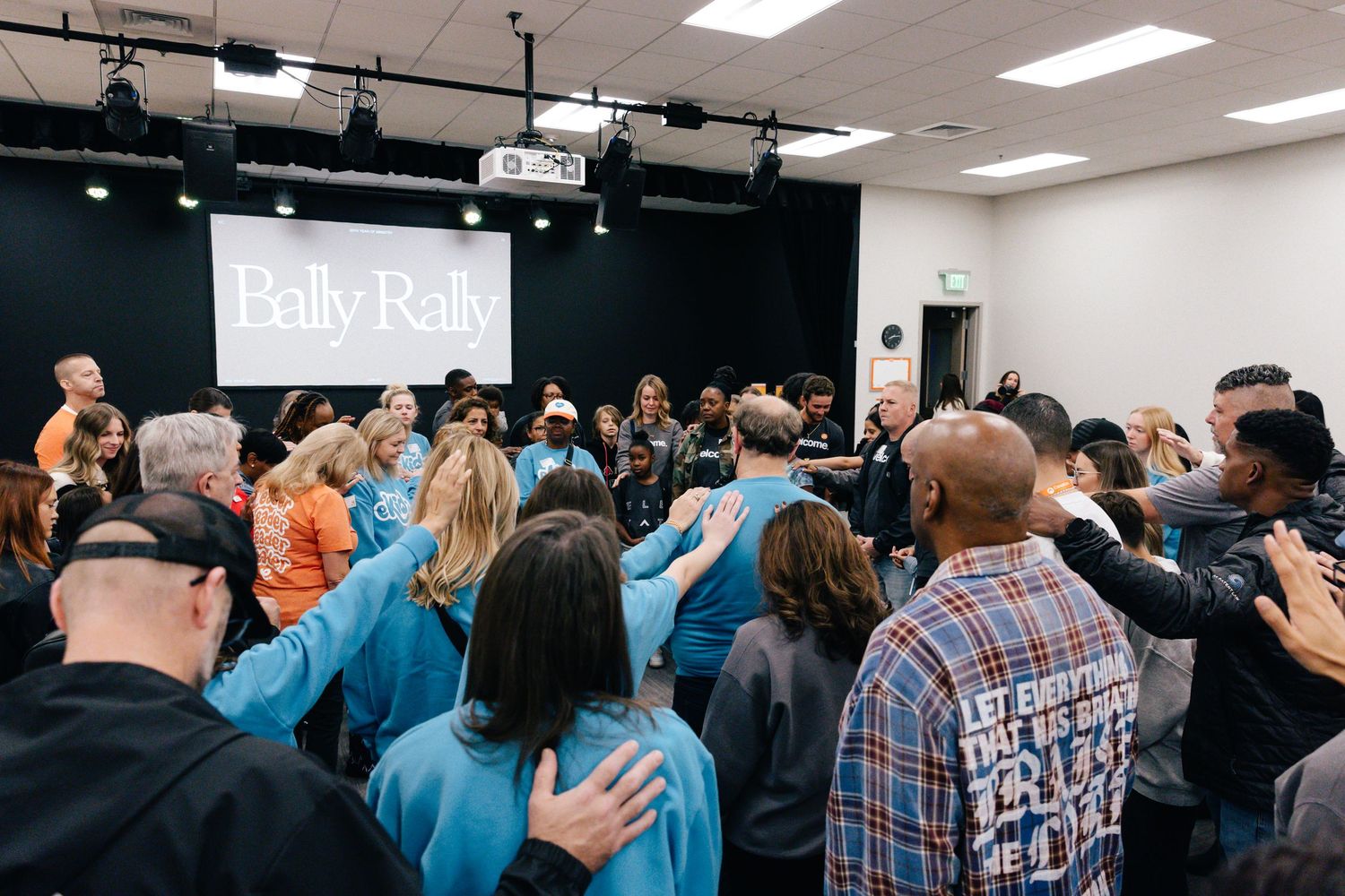 A group of people pray together with hands on shoulders in a room with a Bally Rally sign on stage.