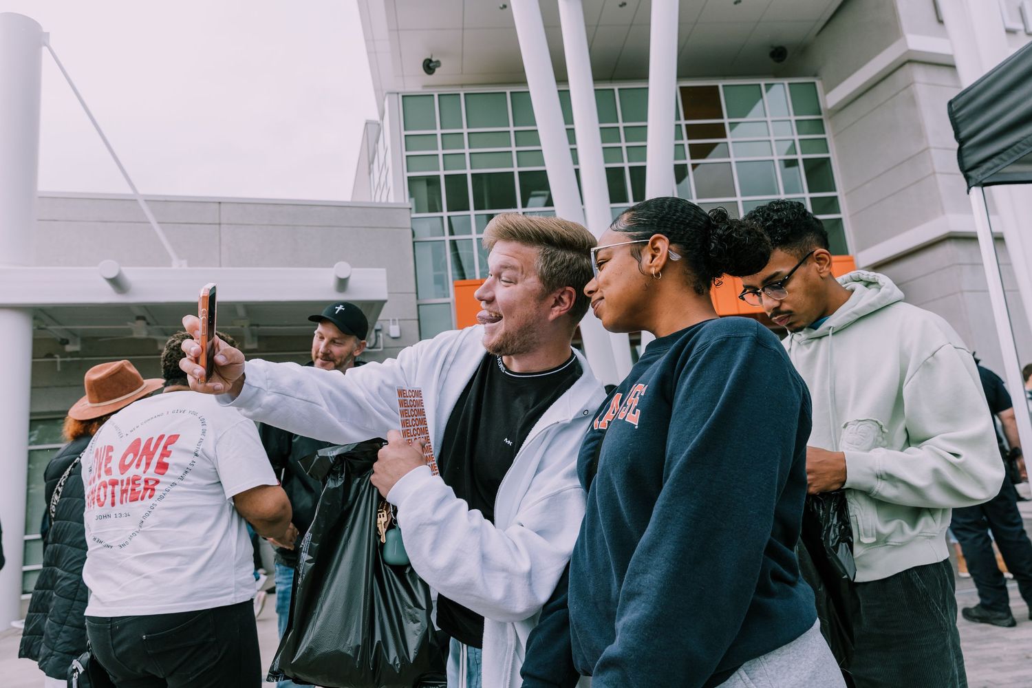 A man takes a selfie with a group of smiling young people outside a modern building at an event.