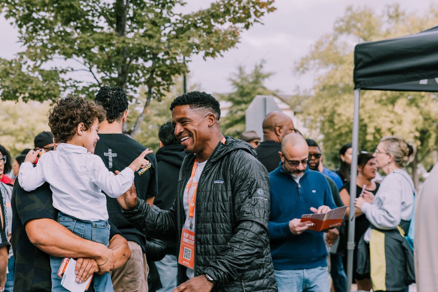 A smiling man laughs joyfully while socializing with others at an outdoor community gathering event.