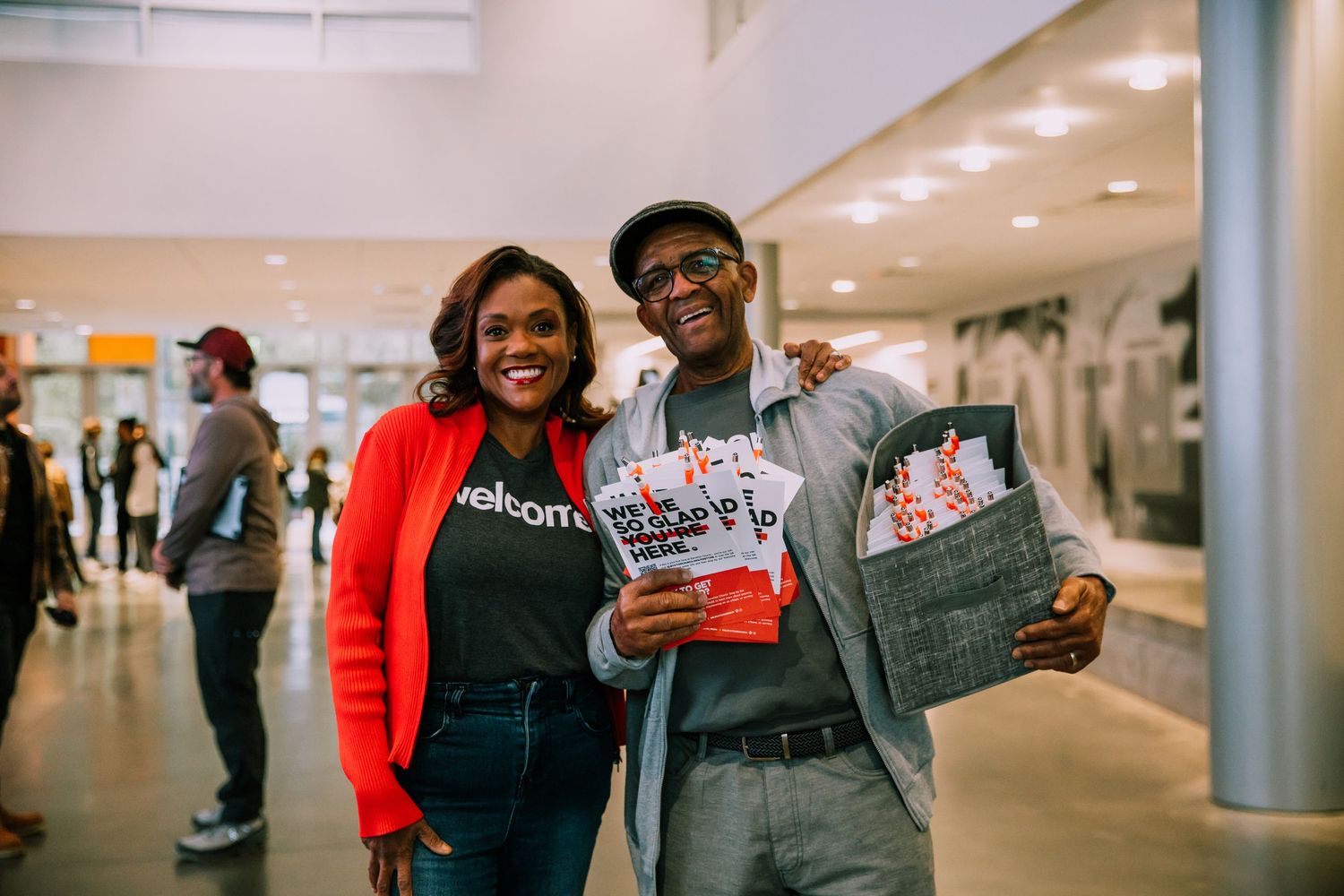 Two smiling people posing together holding welcome bags and materials at a church event.