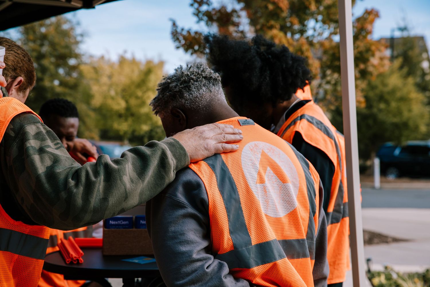 Volunteers in orange safety vests praying together with hands on shoulders outdoors.