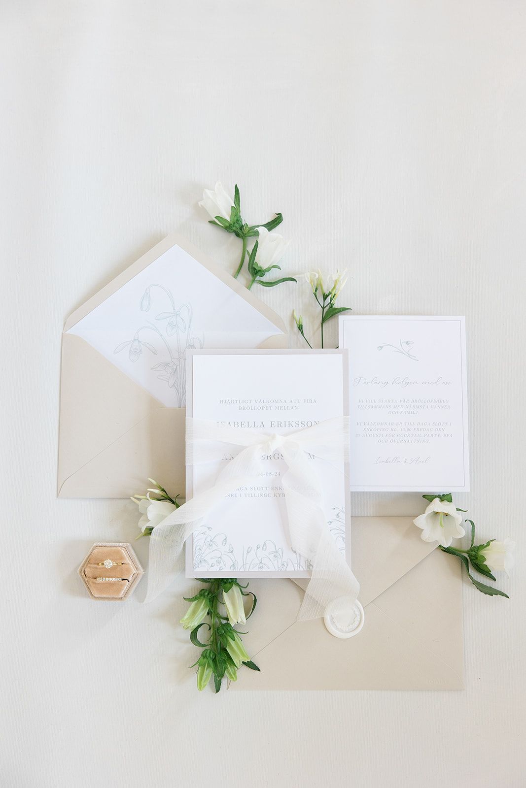 Elegant white wedding invitation suite with beige envelopes and delicate green foliage accents arranged on white surface.