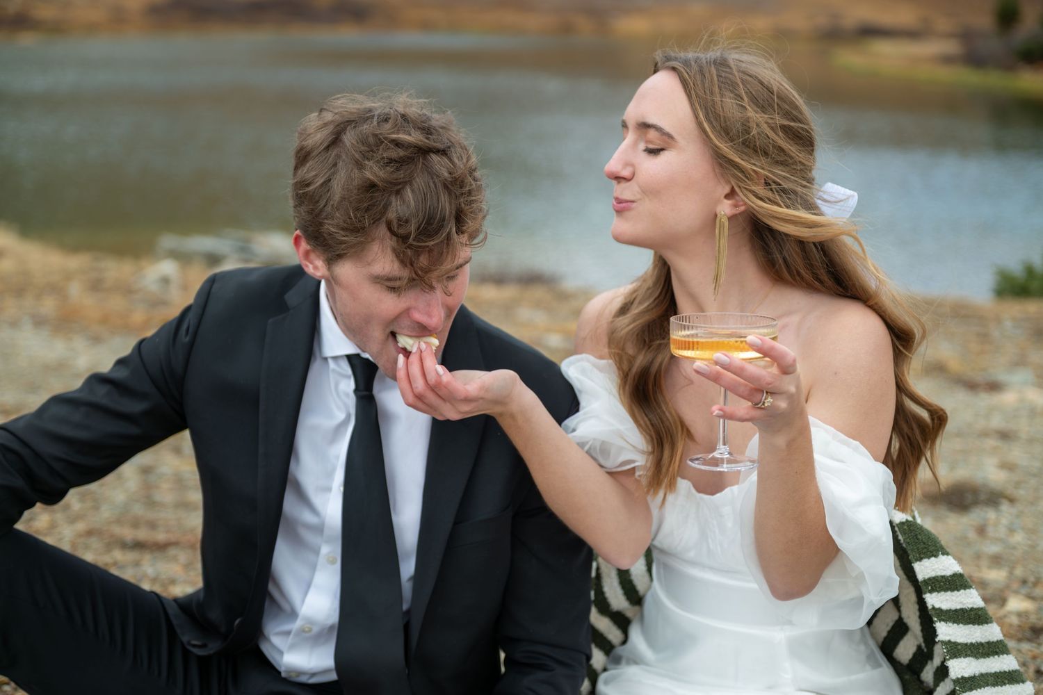 A bride in white off-shoulder gown holds cocktail and feeds her groom in black suit wedding cake by an alpine lake near Breckenridge.