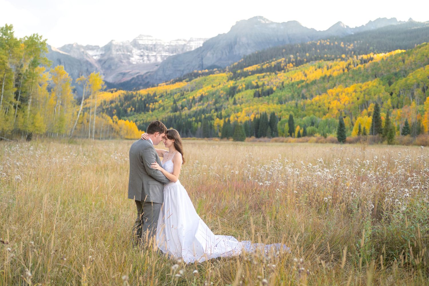 An eloping couple embracing in a mountain meadow during fall foliage, wearing wedding attire with snow-capped peaks behind.