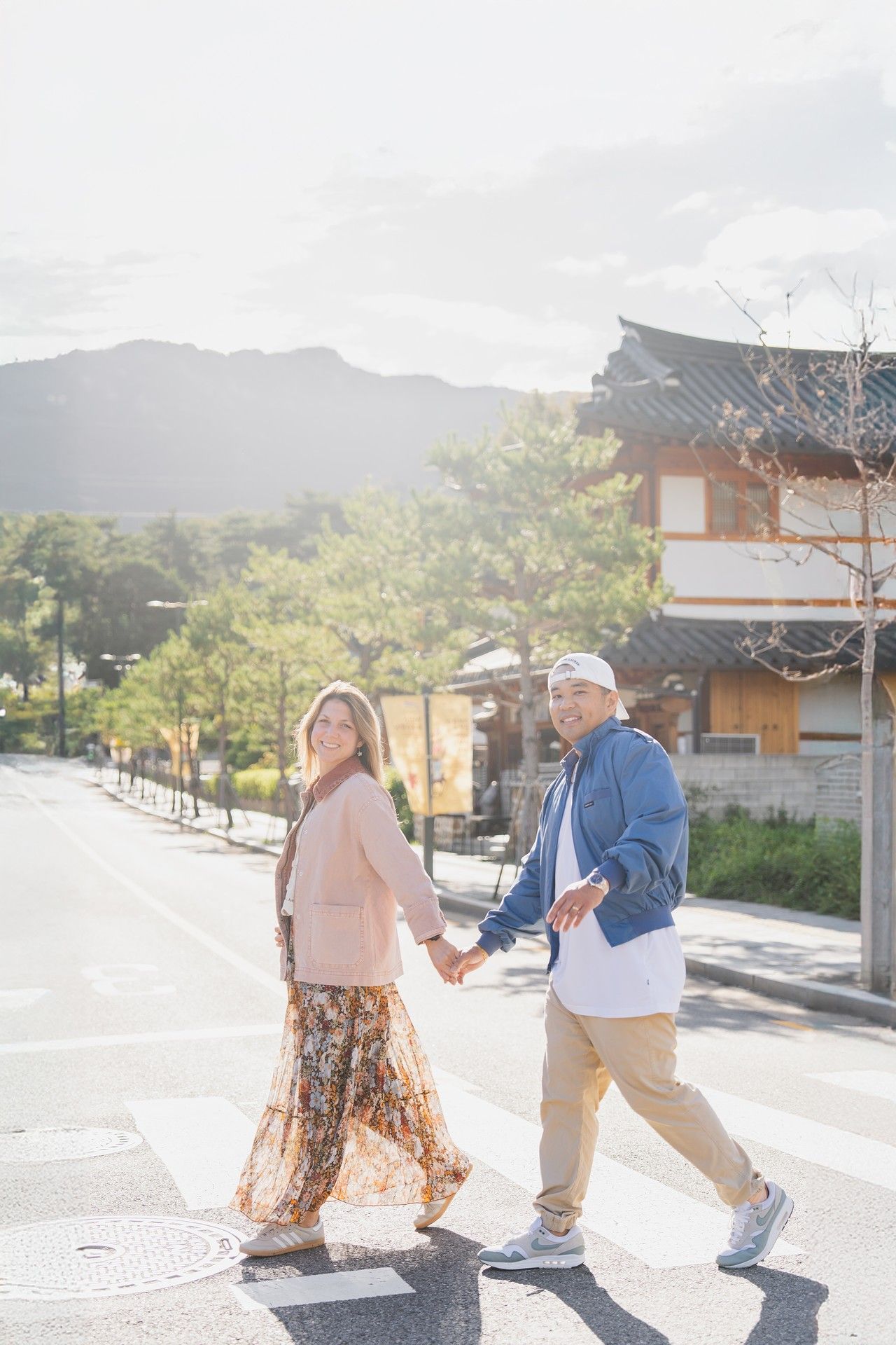 A couple holds hands while walking on a sunny street with traditional Japanese architecture and mountains in the background.