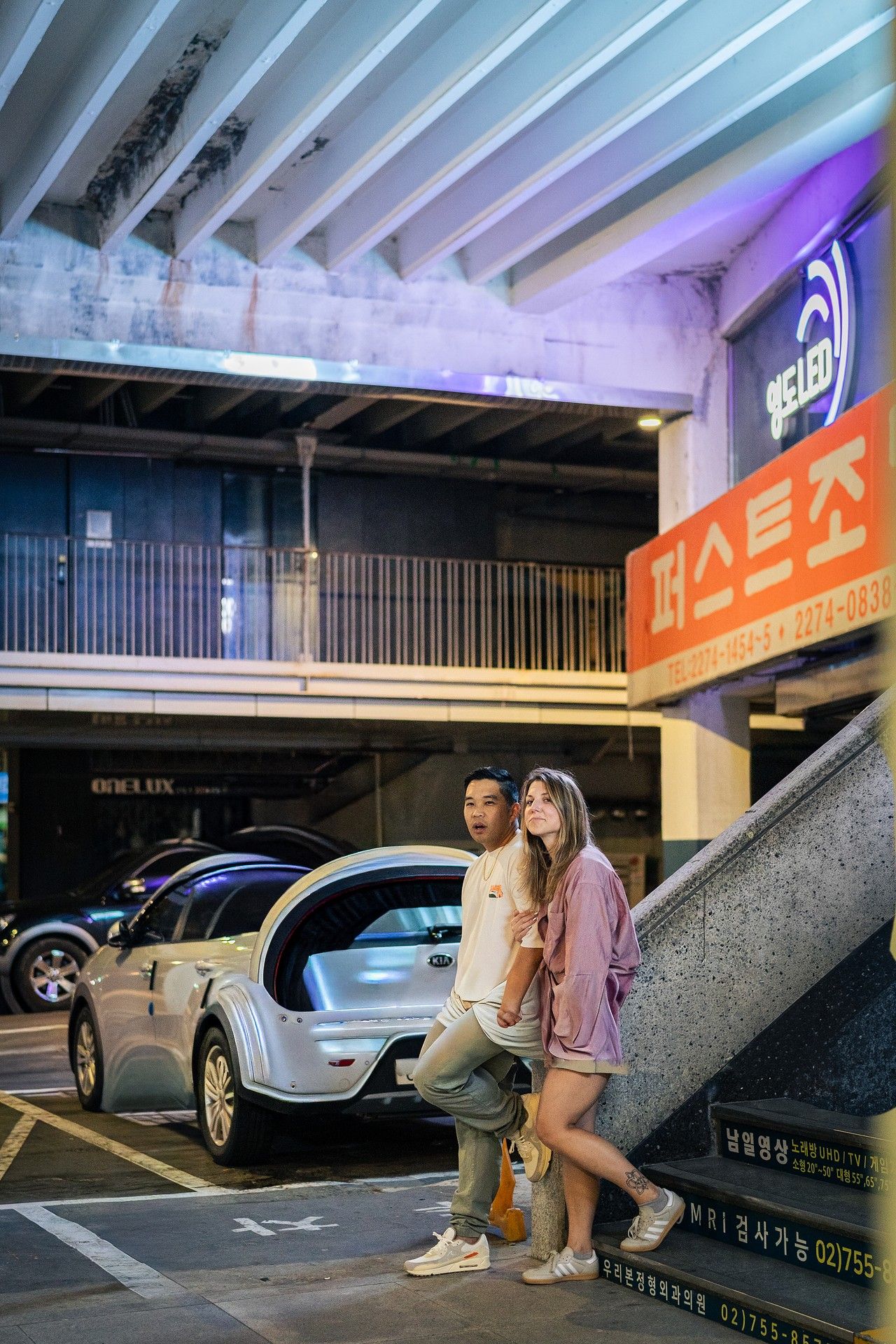 A series of parked cars in an underground parking garage with neon lighting and Korean signage visible.