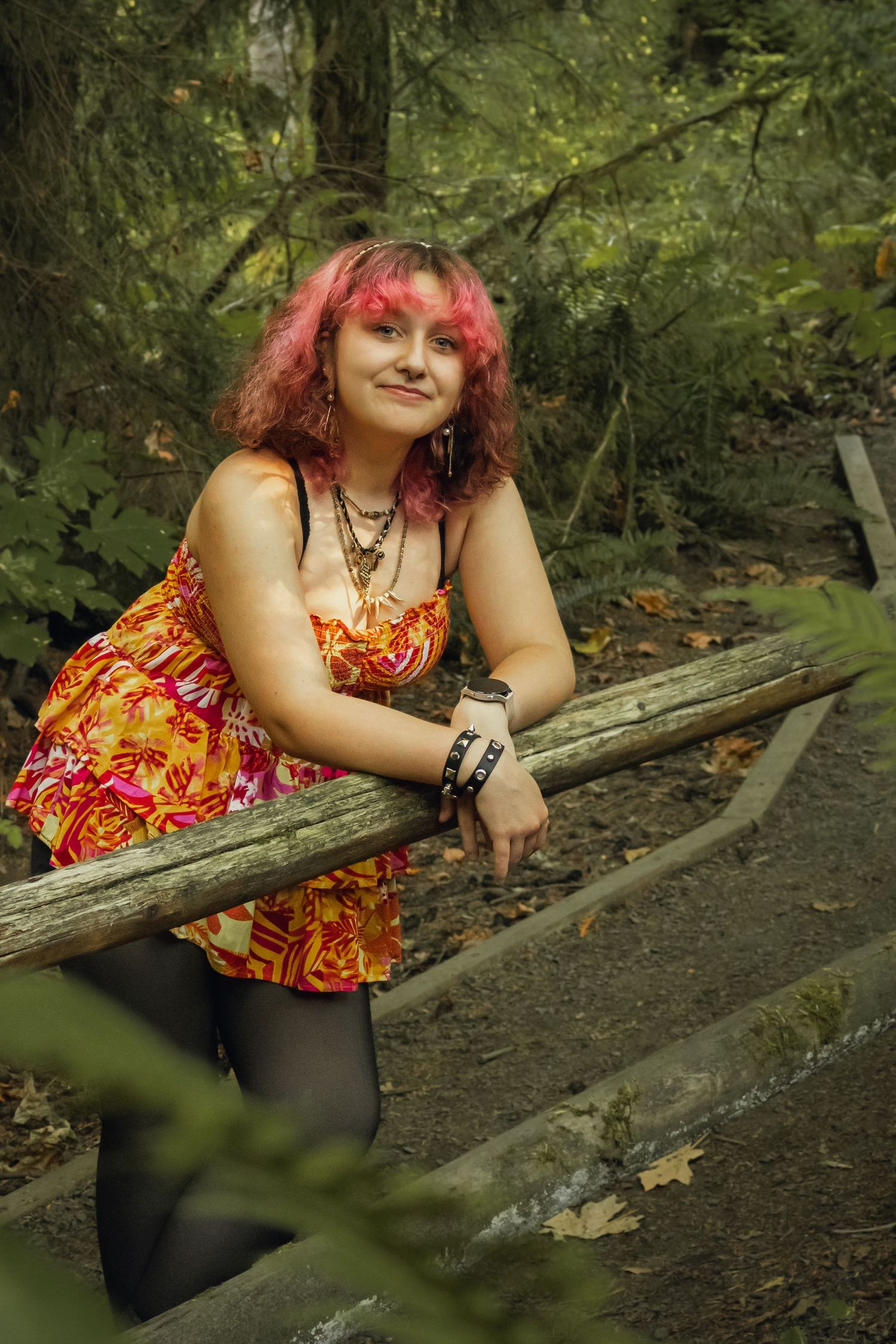 Young woman with pink hair in floral dress leaning on wooden railing in forest.