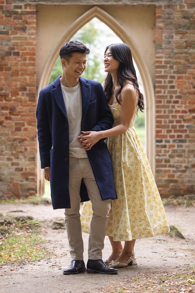A couple in stylish outfits share a laugh under a Gothic archway and brick walls. This is part of an engagement shoot at Painshill Park