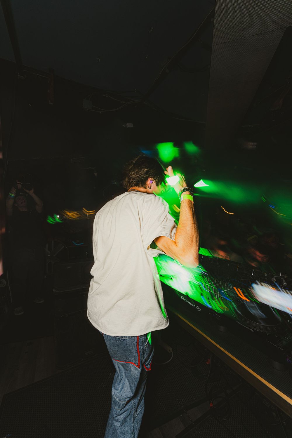 A person in a white shirt and jeans leans forward in a dark room illuminated by green lights.