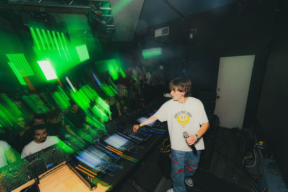 DJ performs at a nightclub with green lighting and electronic equipment on stage.