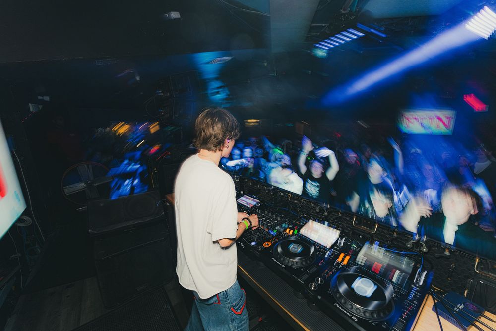 DJ performs at a nightclub with blue lighting and crowd visible in the background.