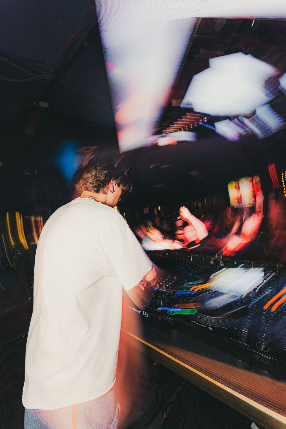 DJ in white shirt performing at a nightclub mixing desk with blurred motion effects in dark atmospheric lighting.