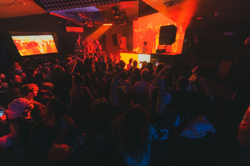 Packed dance floor bathed in red lighting with projection screens and crowd facing the performance area.