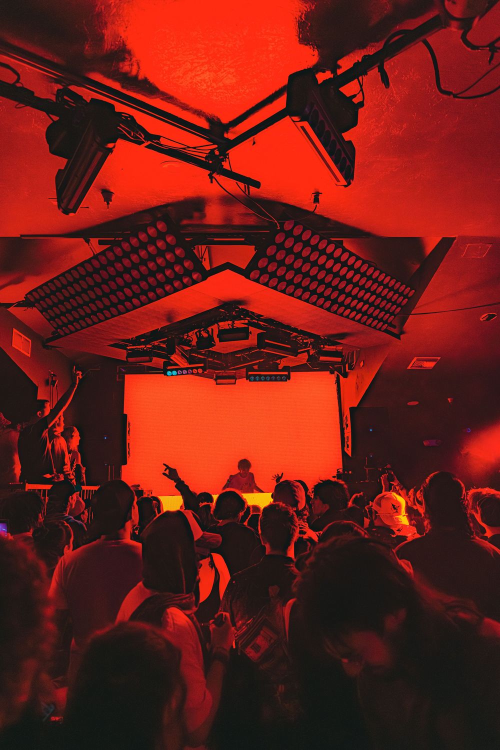 A crowded nightclub dance floor bathed in red lighting with stage equipment and speakers mounted overhead.