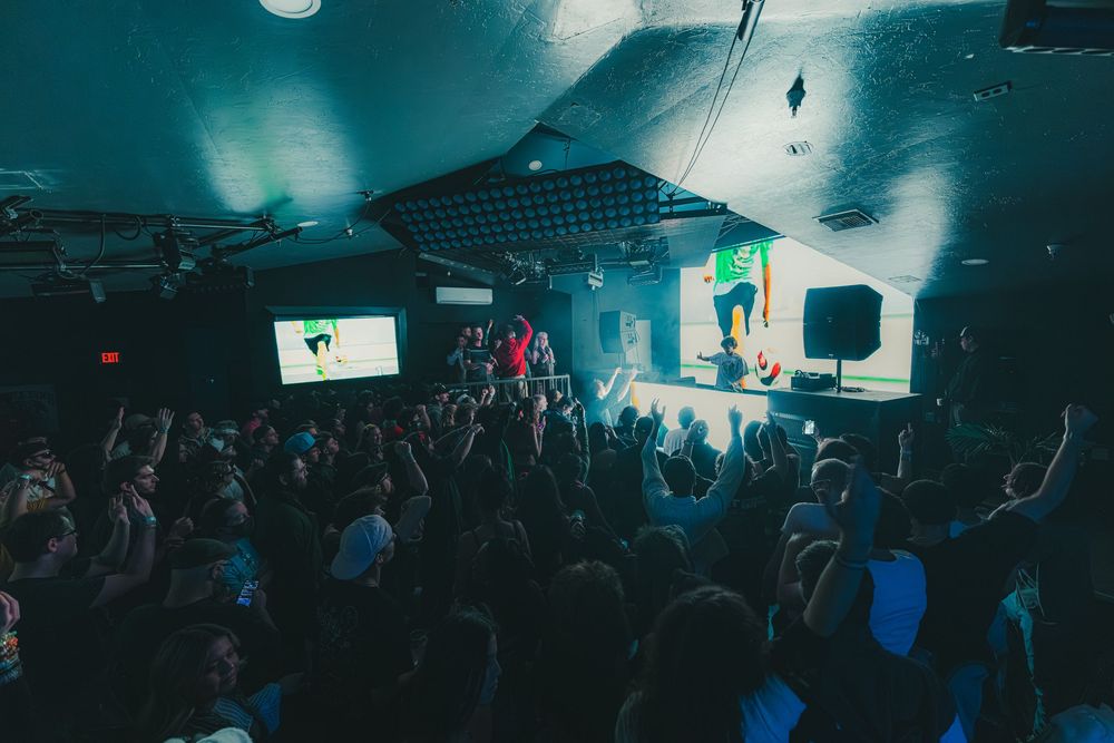 A dark nightclub interior with blue lighting and silhouettes of people dancing and socializing on the dance floor.