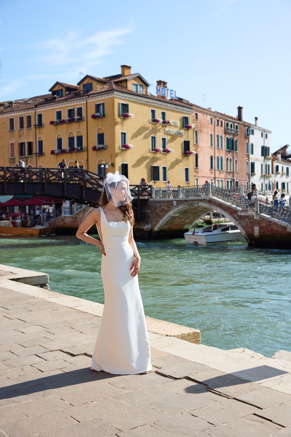 Person in white dress walking along Venice canal with historic buildings and bridge in background.