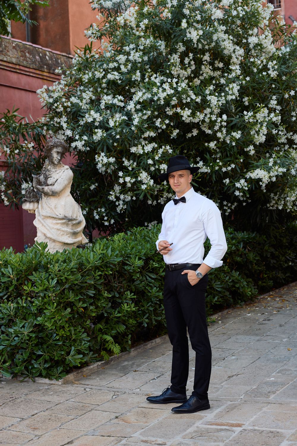 Wedding couple poses in formal attire near blooming white flowers and garden statues.