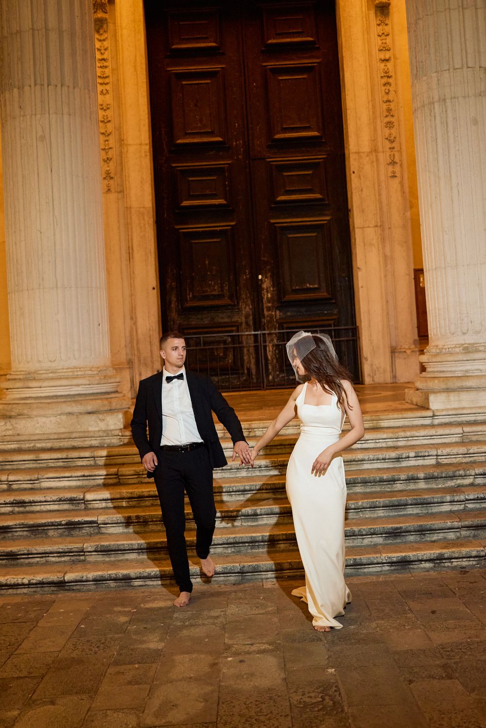 Elegant couple walking along ornate stone steps of historic building at night.