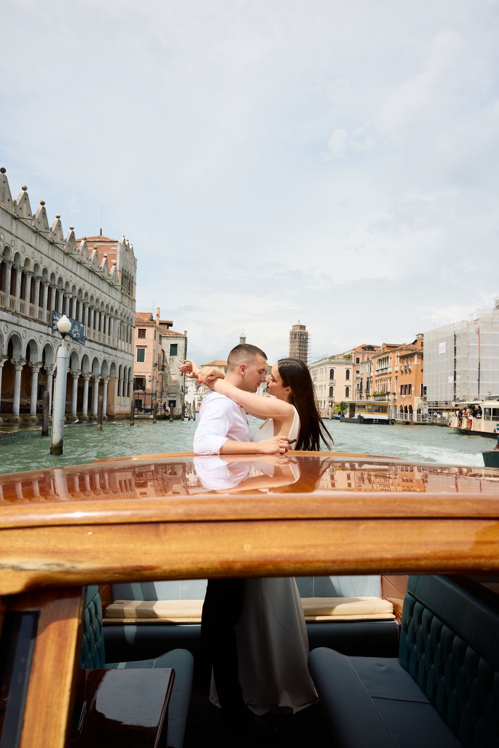A romantic couple lying on a wooden boat deck in Venice with historic buildings and canals in the background.