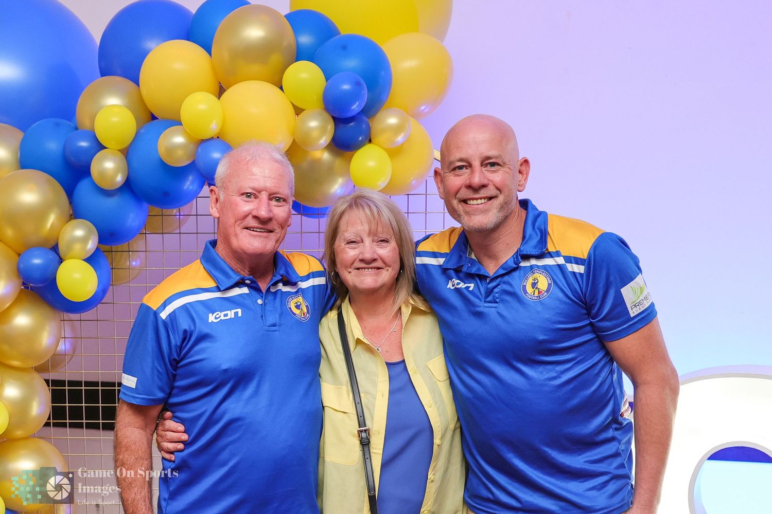 Three people in blue and yellow team attire pose together at an event with colorful balloon decorations.