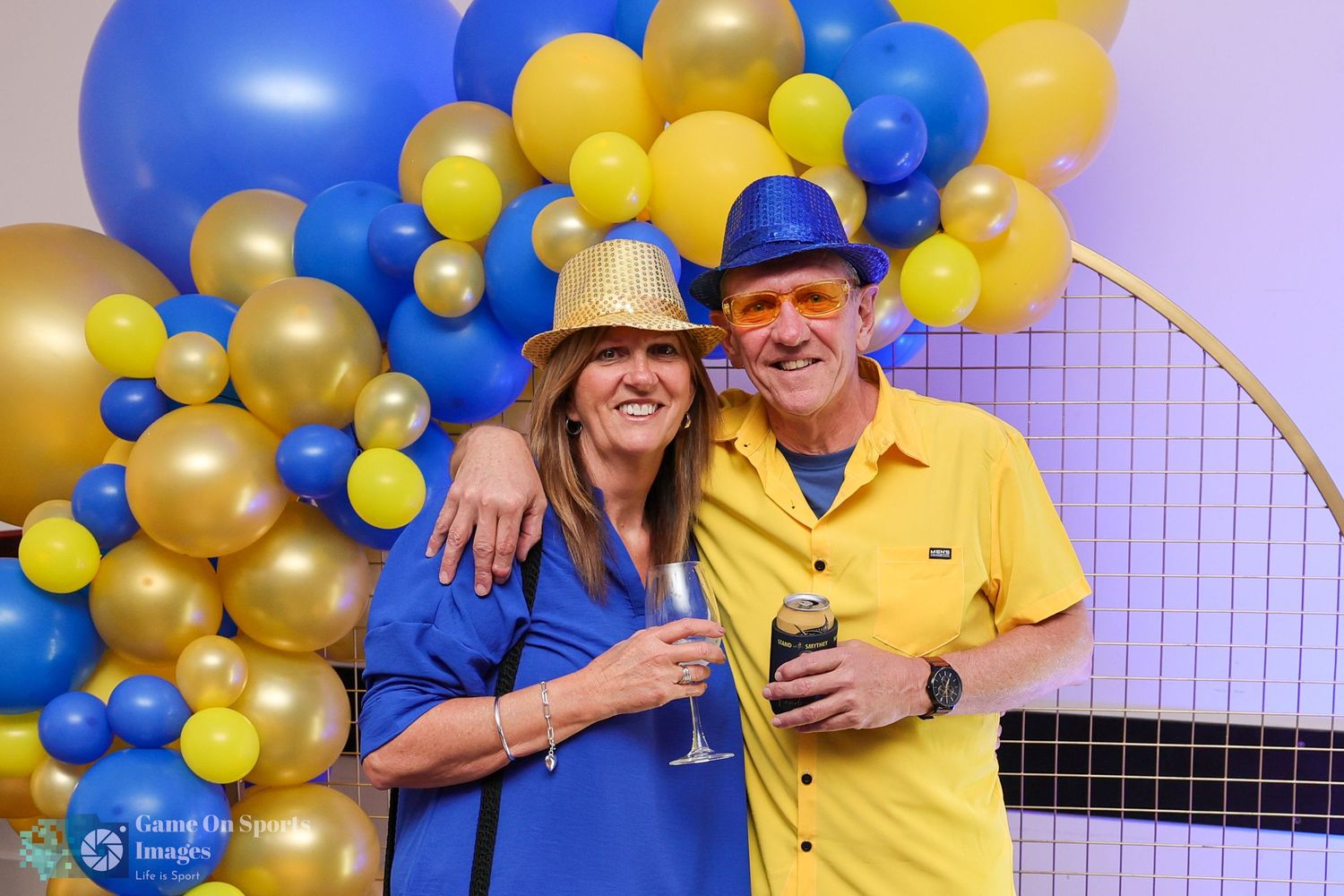 Two people in blue and yellow outfits pose together in front of a festive balloon arch with gold and blue balloons.