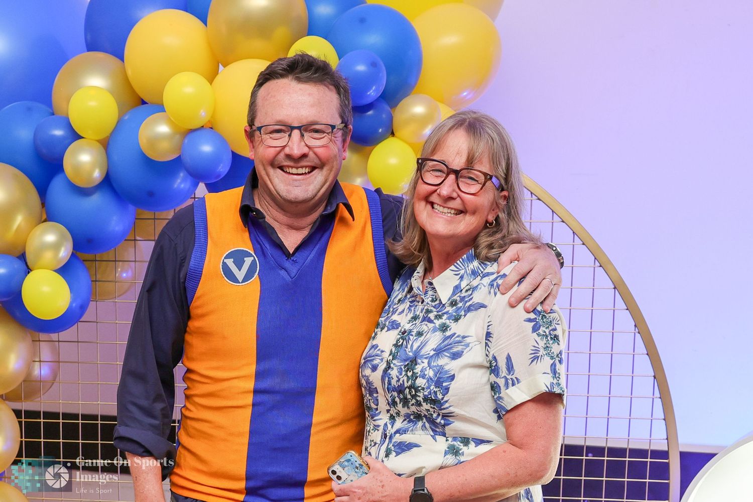 A couple poses together at a celebration decorated with blue and yellow balloons in the background.