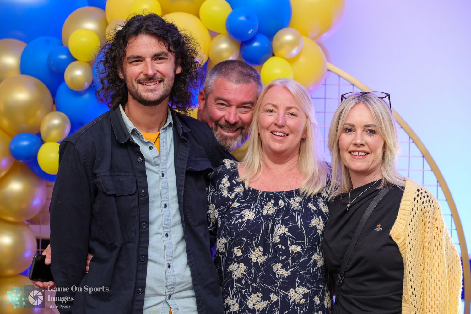 A group poses together at a celebratory event with gold and blue balloons decorating the background.