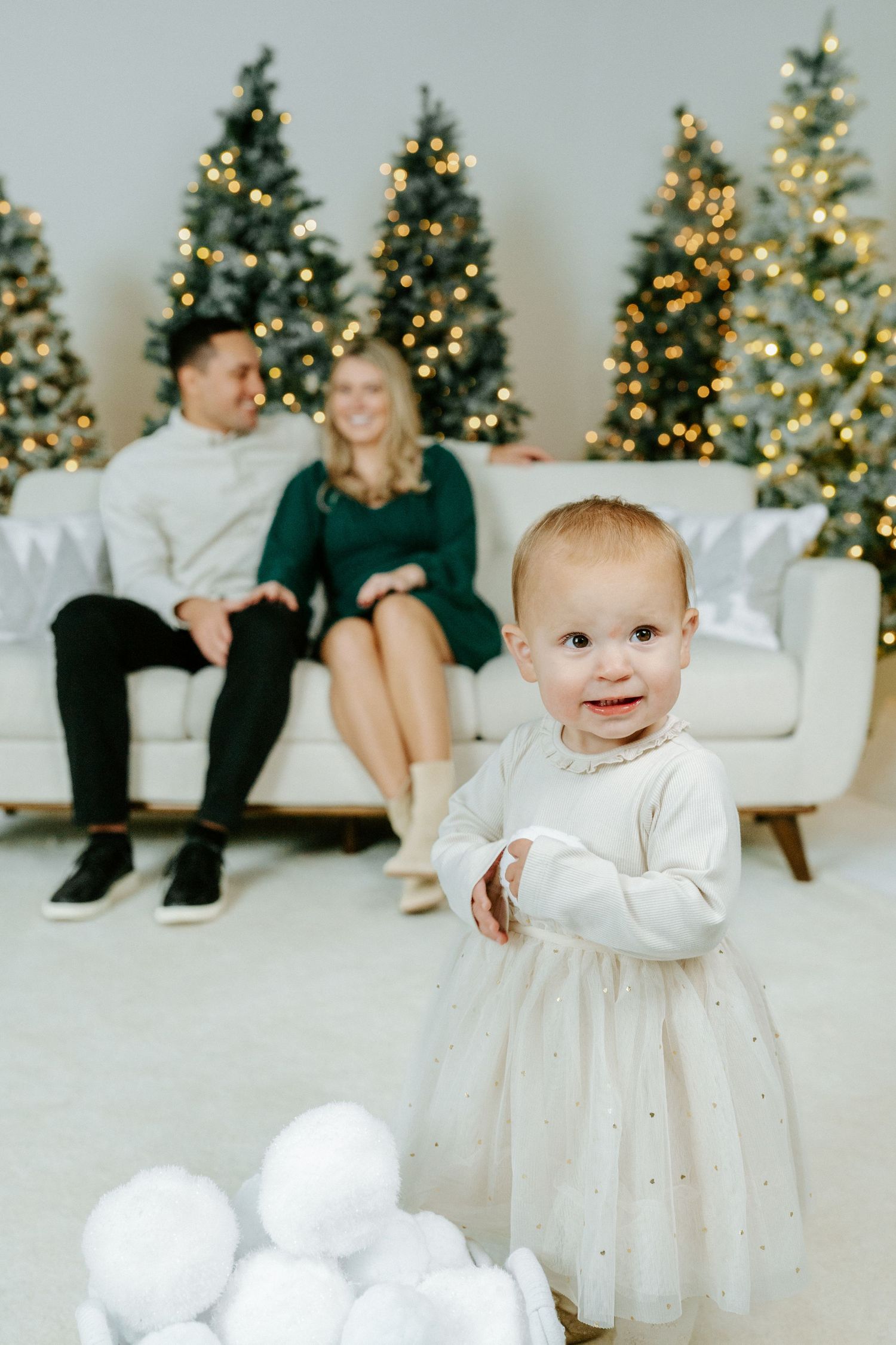 A baby in a white dress smiles while standing in front of multiple decorated Christmas trees in a bright studio setting.