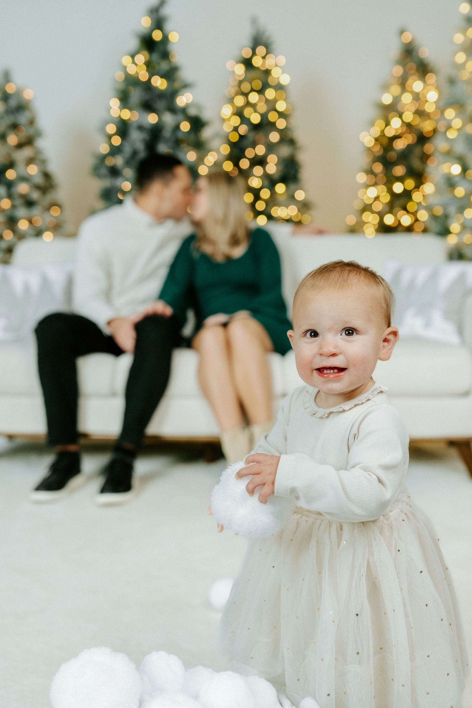Baby in white sparkly holiday dress smiles and crawls on white floor with decorated Christmas trees in background.
