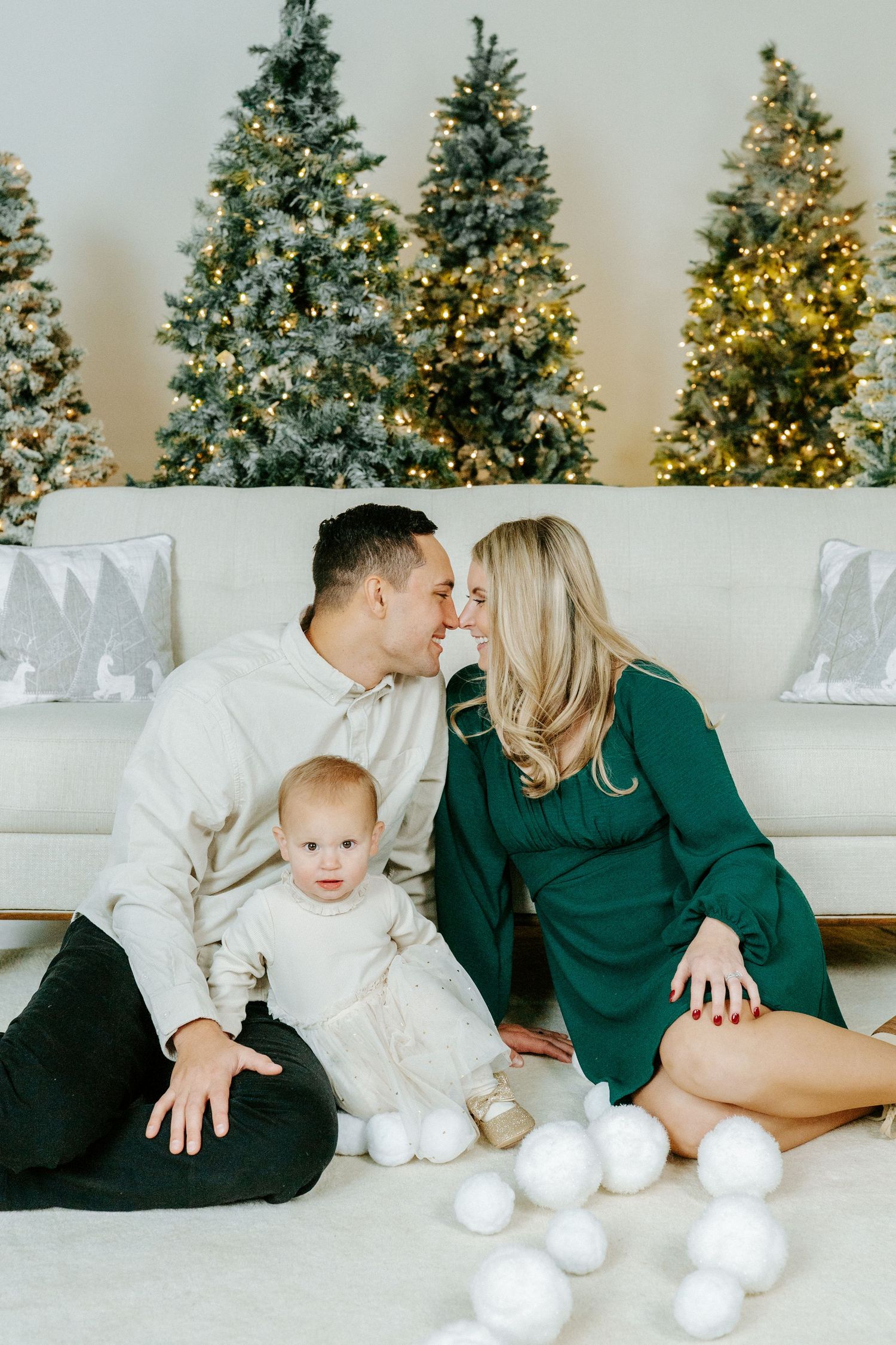 A cozy holiday scene with a couple and baby sitting on white couch in front of decorated Christmas trees.