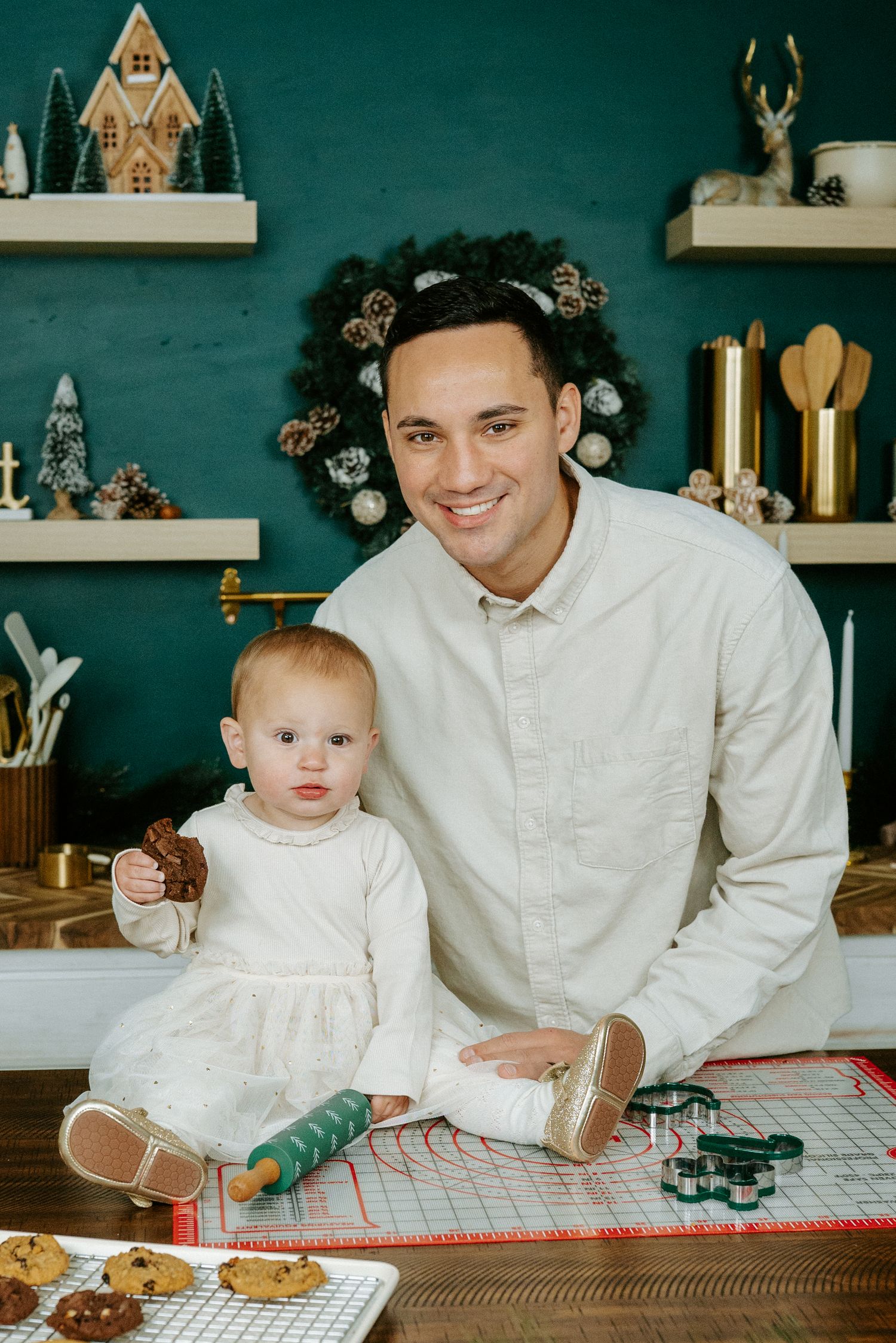 A parent and baby sit together at a wooden table with toys and decorative items on shelves in the background.