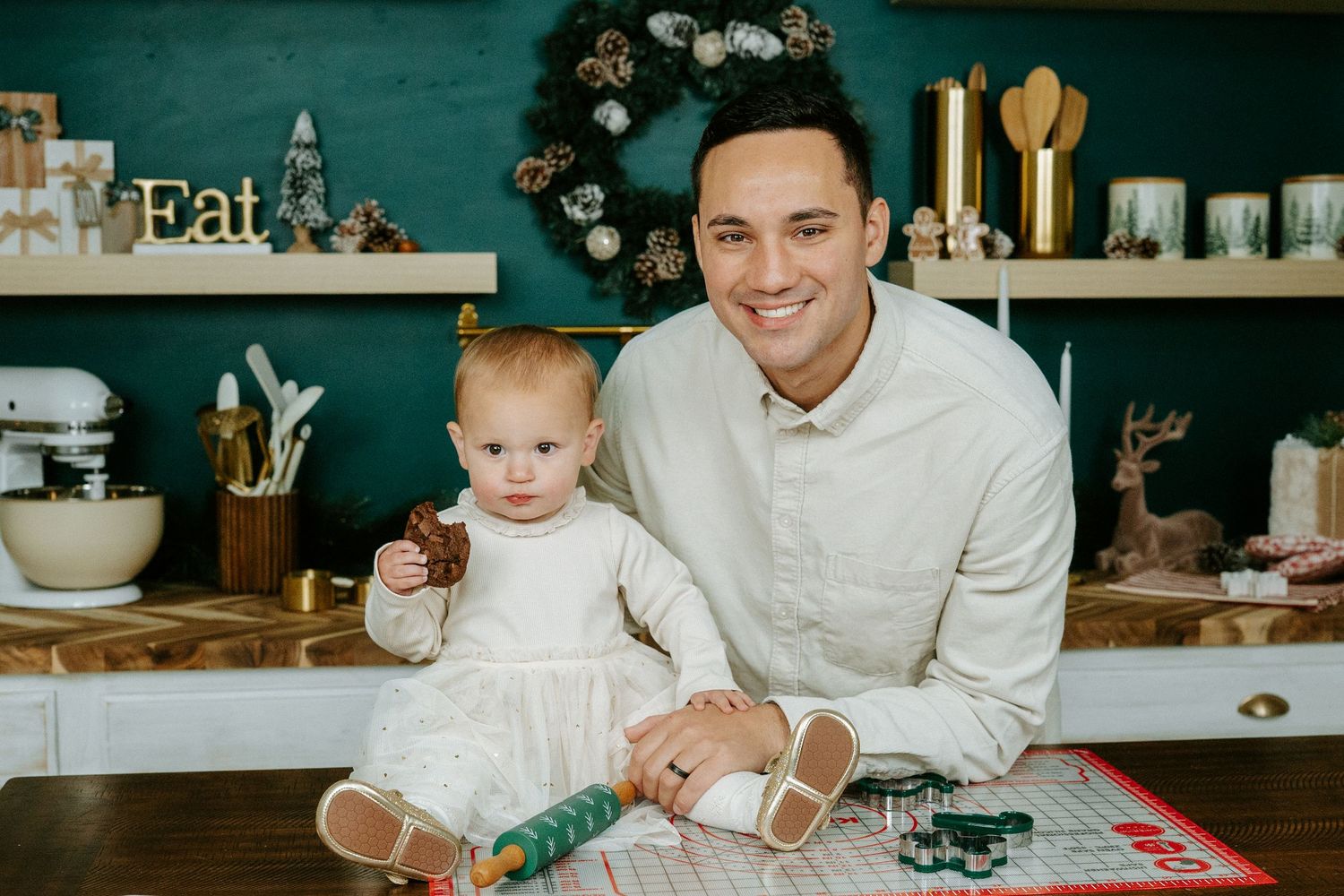 A cozy kitchen scene with a white-clad pair sitting at a counter with holiday decorations and cooking items in the background.