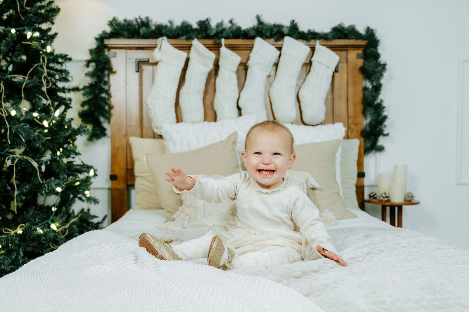 A baby sits in a white bed surrounded by Christmas decorations and twinkling lights.