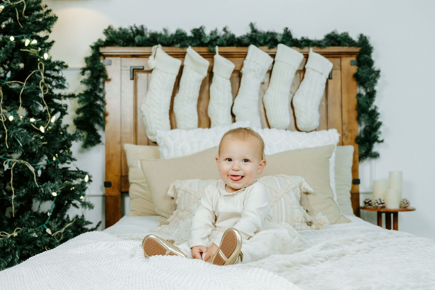 A baby wearing white clothing sits on a white bed with pillows and a Christmas tree in the background.