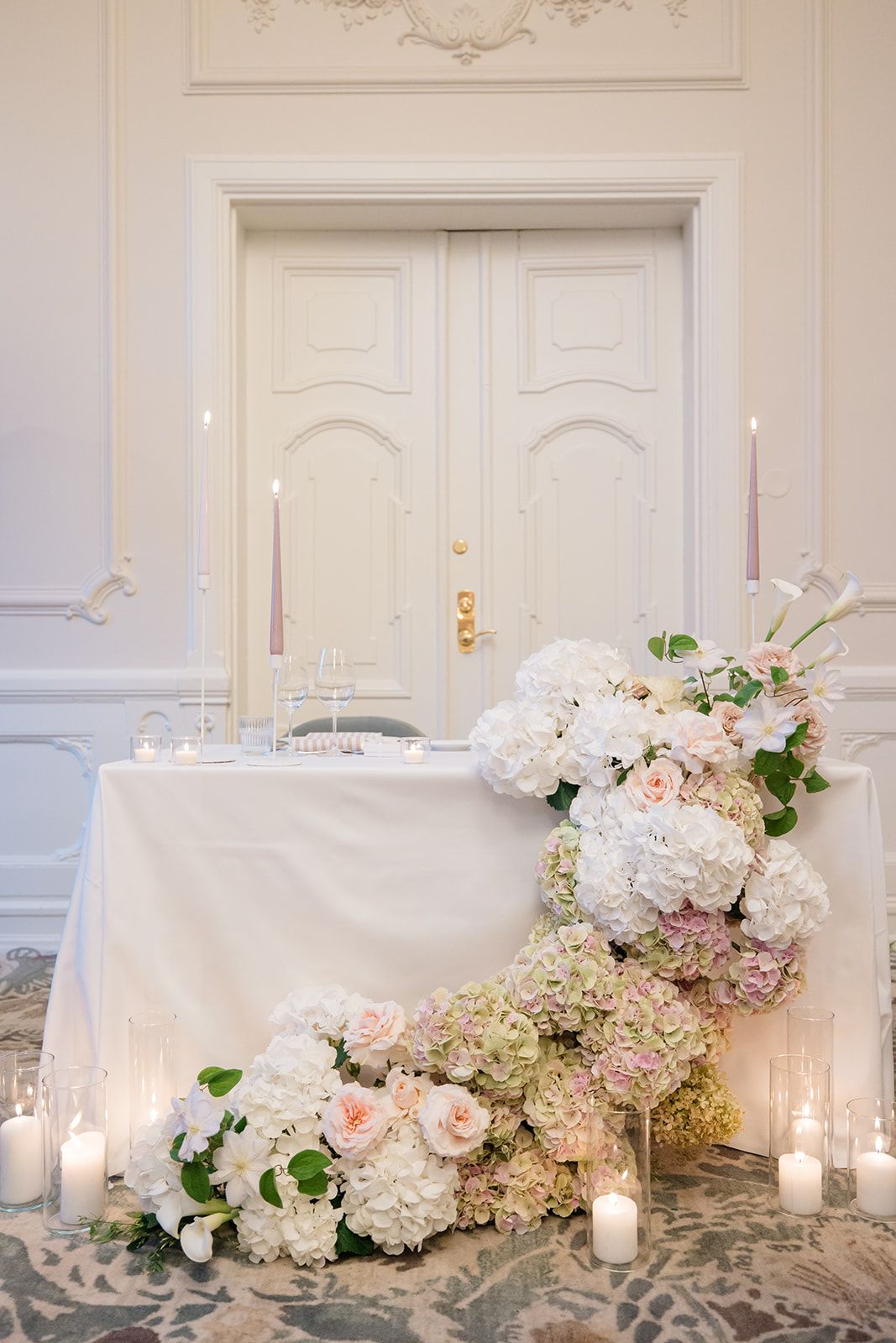 Elegant white floral arrangements with hydrangeas and roses adorn a wedding reception table with glowing candlelight.