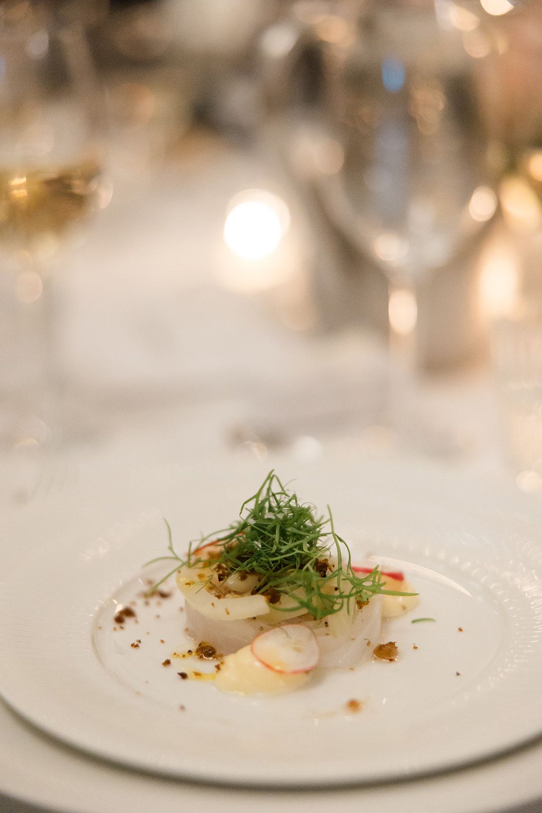 Elegant plated dish with delicate microgreens and garnishes on a white plate with soft bokeh lighting in background.