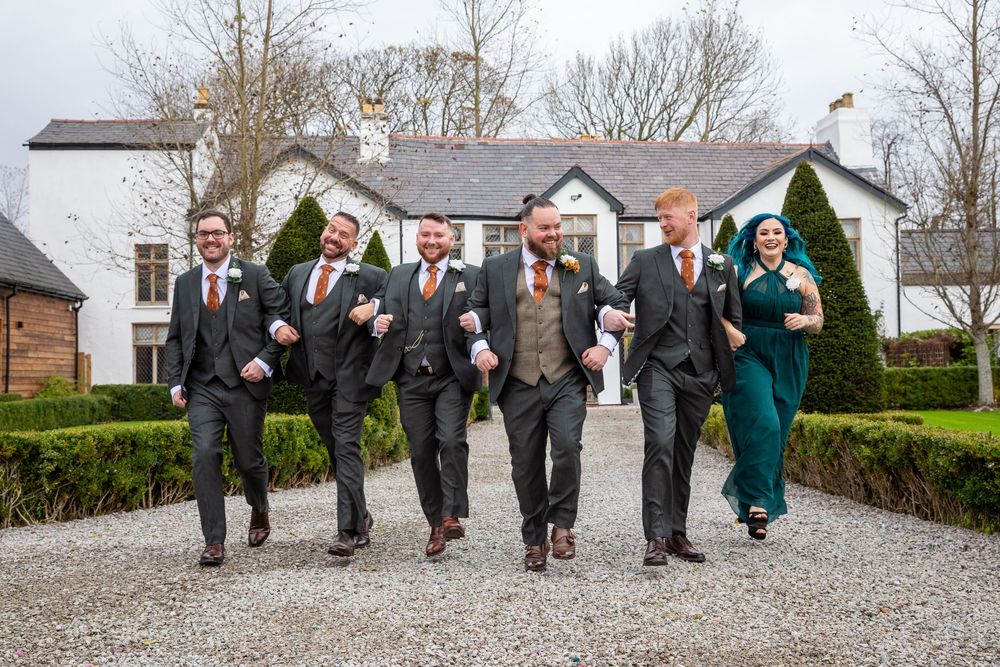 Groom and groomsmen in grey suits walk confidently along a gravel path outside a wedding venue.