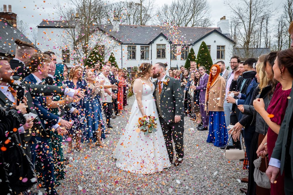 Bride and groom kiss as colorful confetti fills the air surrounded by cheering wedding guests outside.