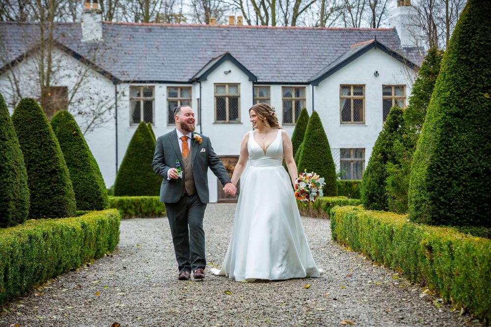 Bride and groom walking hand in hand along a gravel path through a manicured garden with topiary hedges.