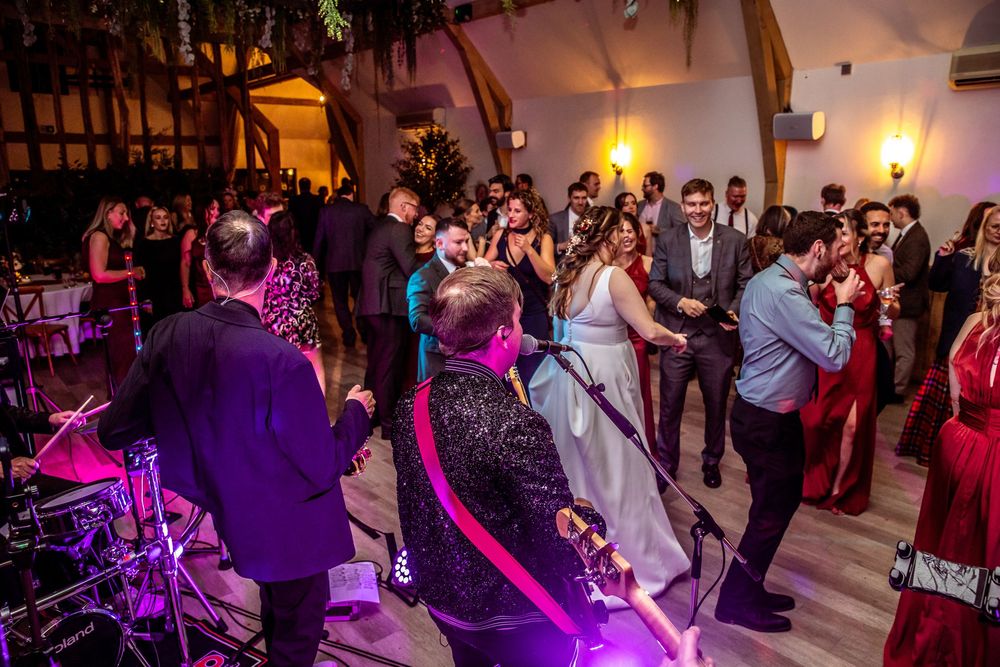 Bride and groom dancing energetically with guests on a crowded wedding reception dance floor with live band.