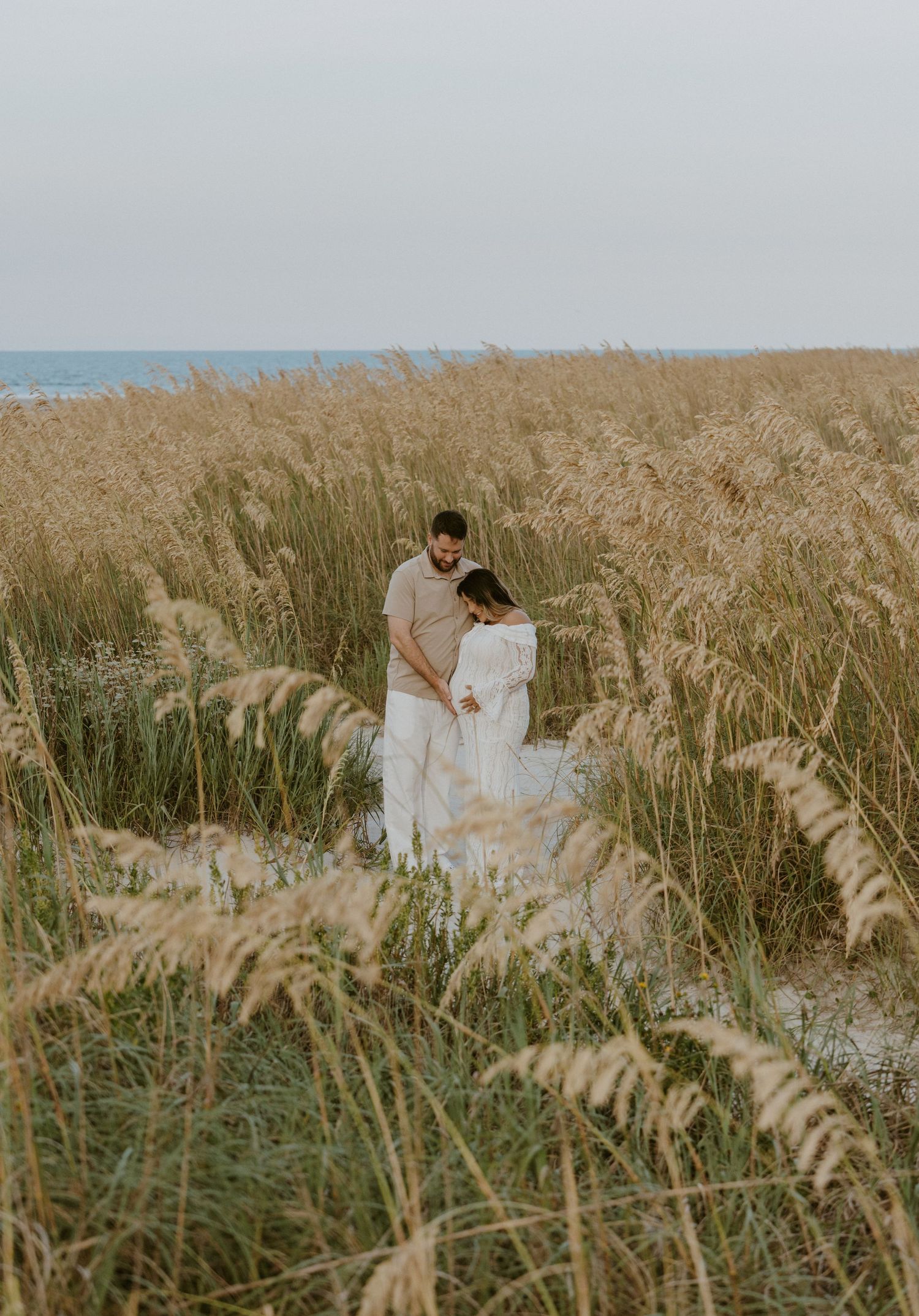 A couple in white attire walks along a sandy beach path at sunset with palm trees in the background.