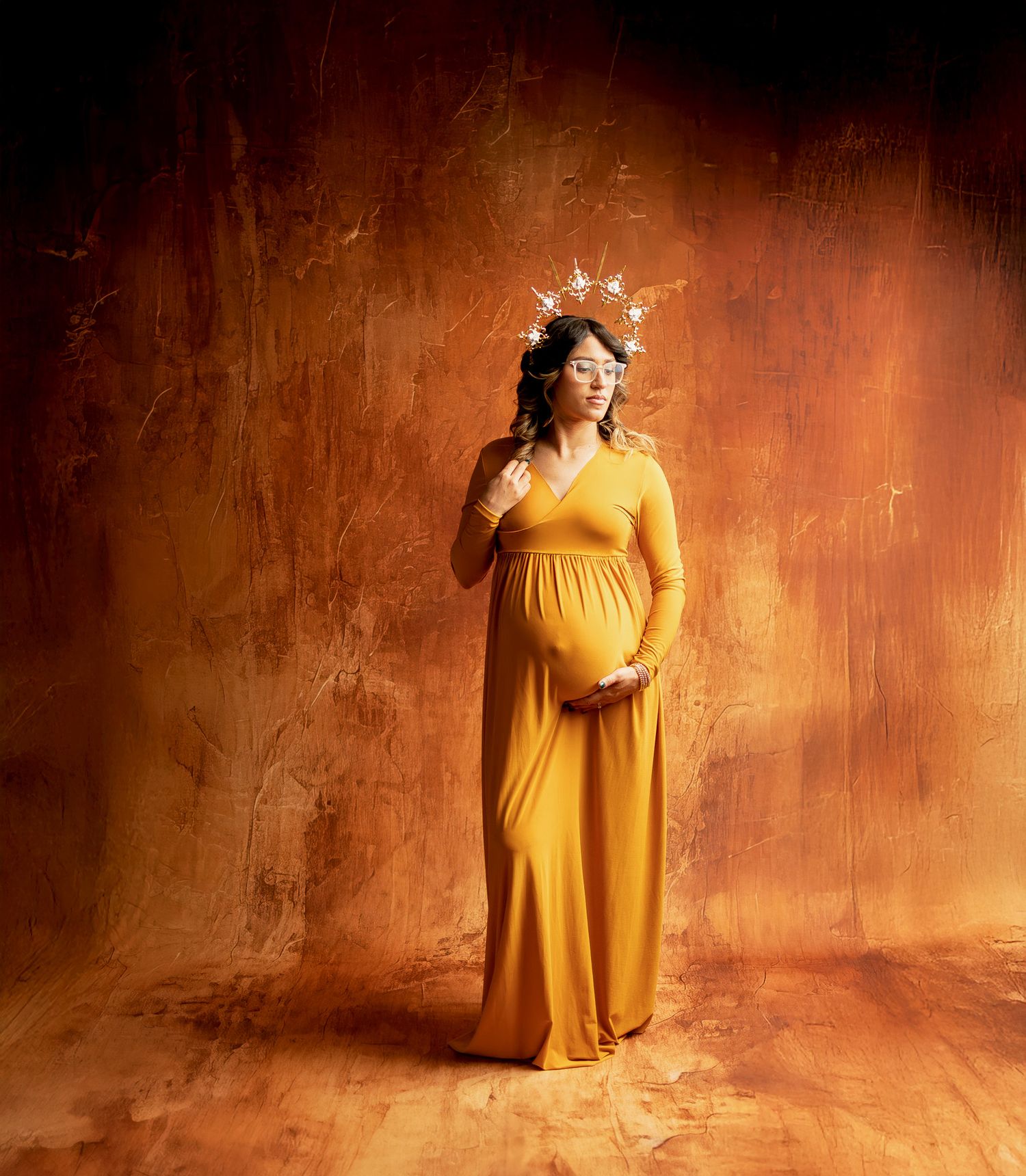 A pregnant woman in a flowing yellow dress and floral crown poses against a warm rustic backdrop with dramatic lighting.