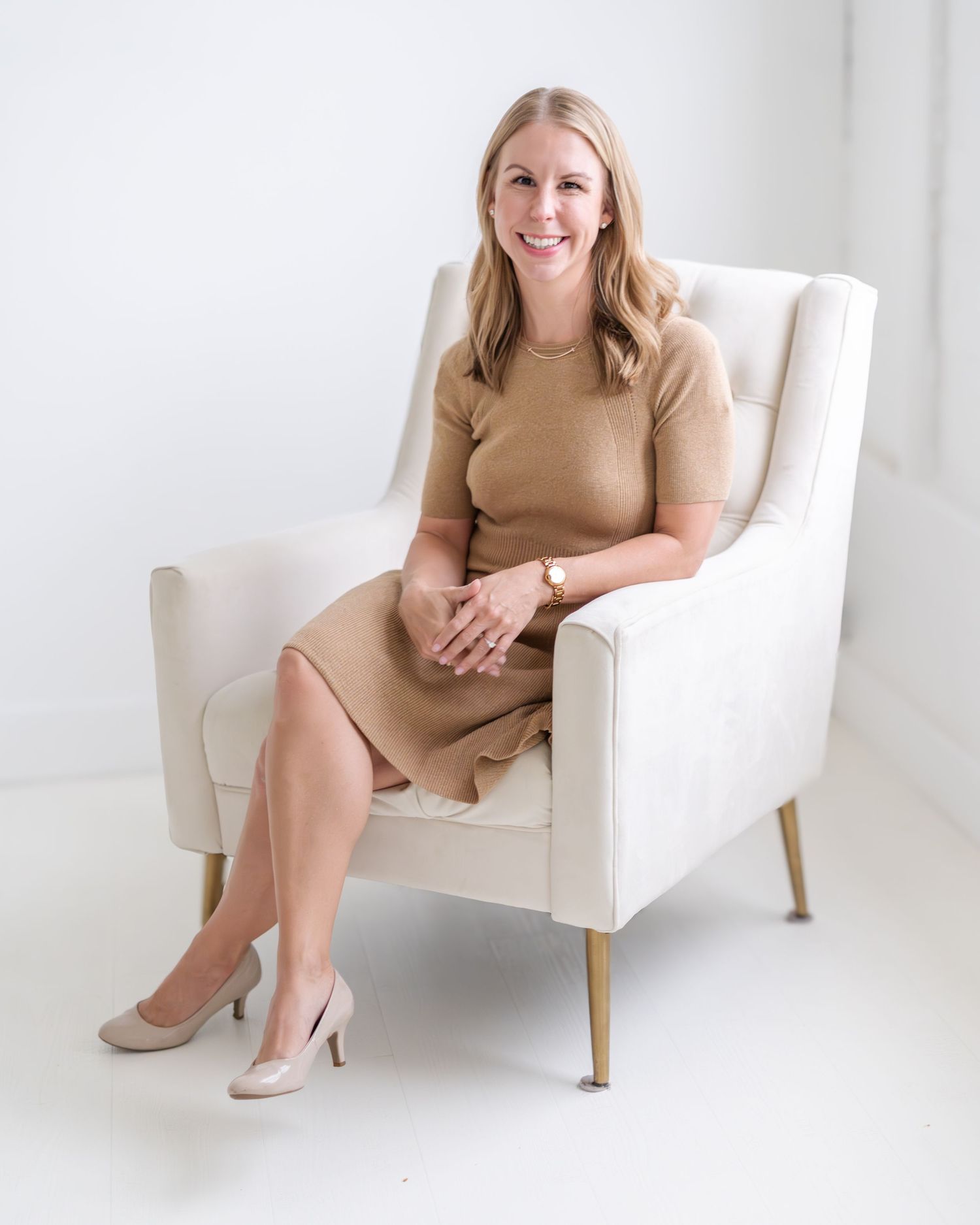 A person in a beige dress sits in a white armchair in a bright minimalist studio setting.