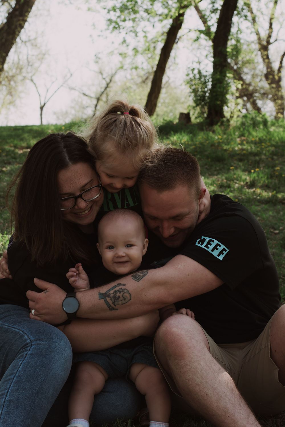 Intimate family cuddle session captured in a wooded outdoor setting with natural lighting.