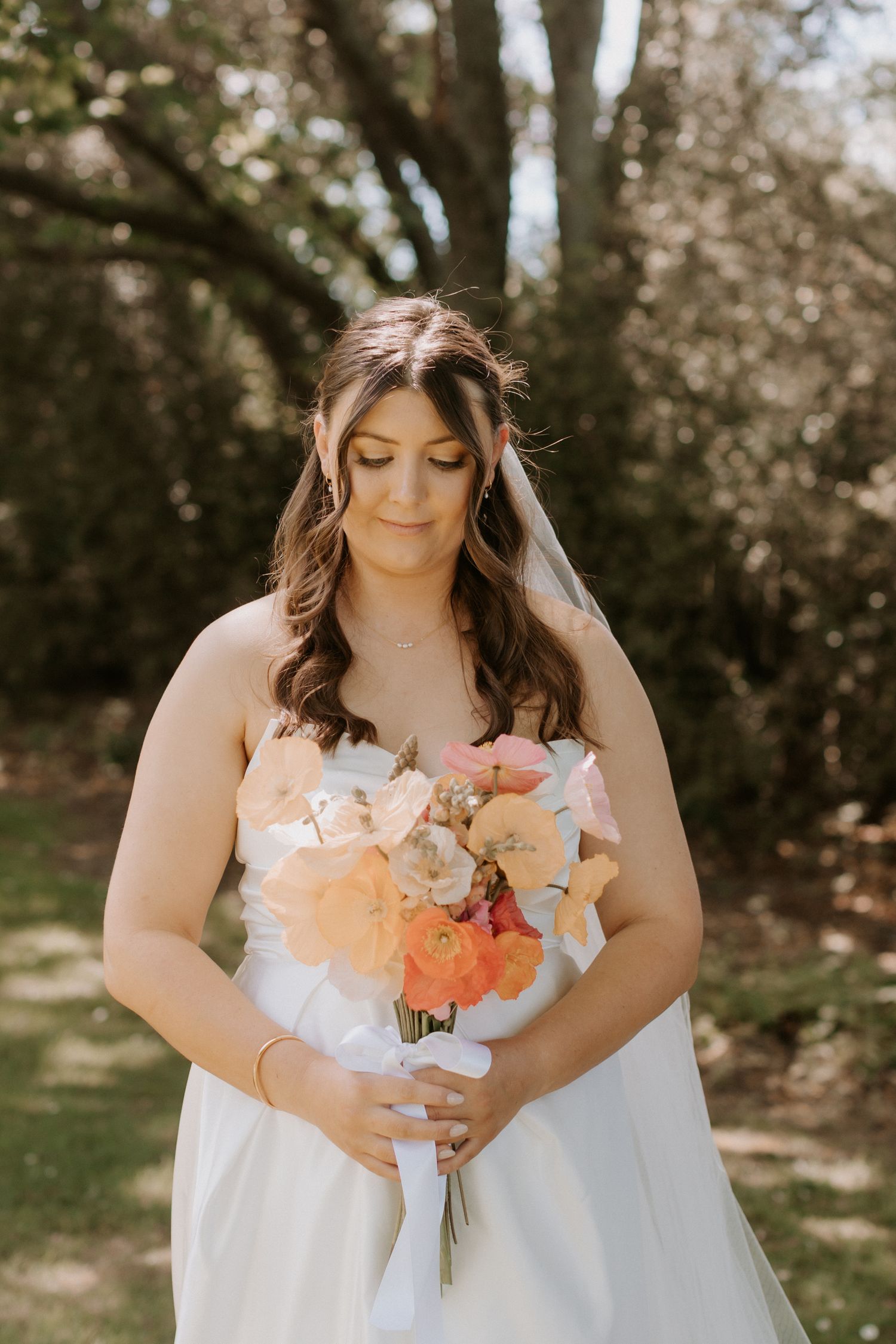 A bride in a white dress holds a peach and coral bouquet while standing in a sunlit garden setting.