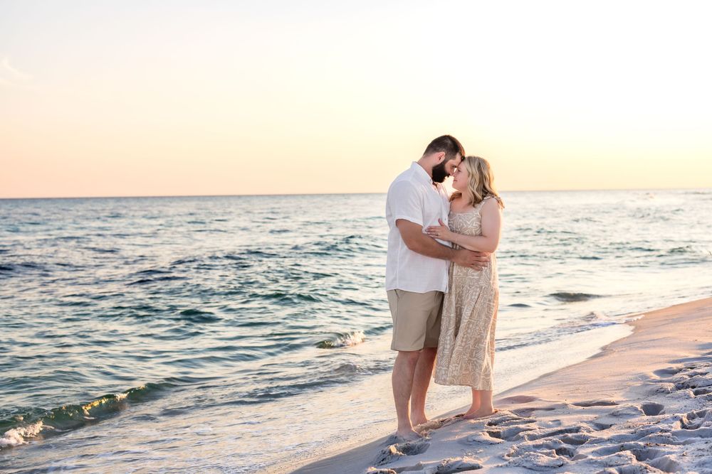Romantic couple embraces on sandy beach at sunset with gentle waves and golden light in the background.