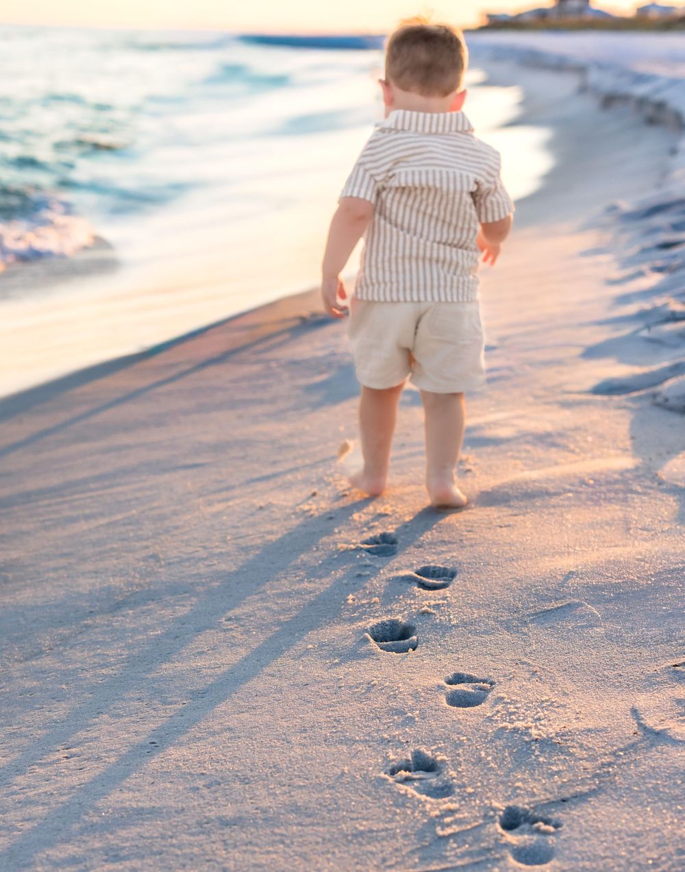 Young child in striped shirt walks along beach leaving footprints in sand during golden hour.