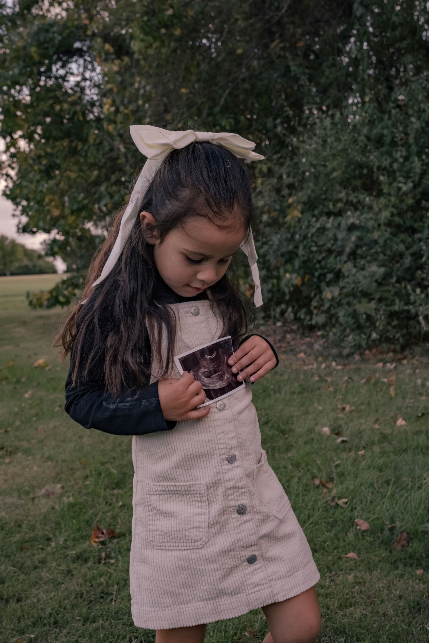 A young child in a beige button-up dress plays outdoors near trees and grass.